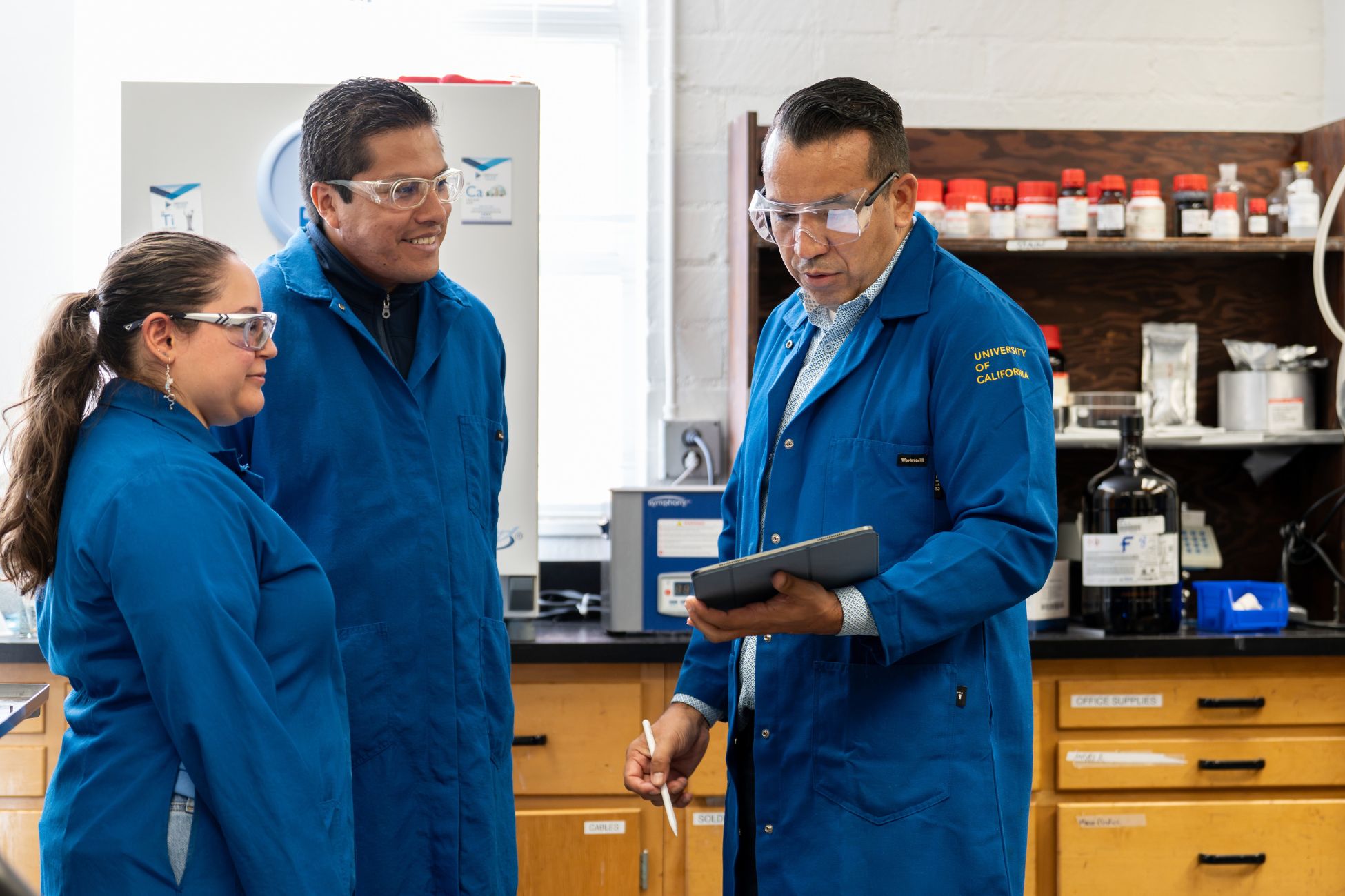 Three individuals in lab coats examining a tablet together, engaged in a discussion about their findings.